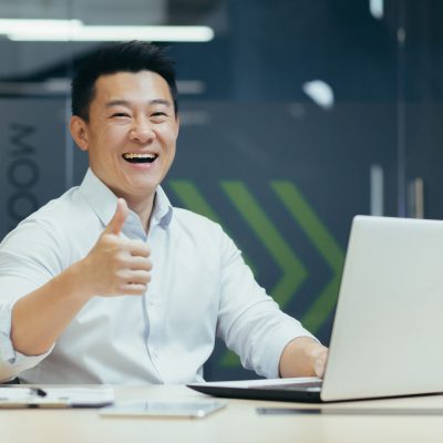 Successful asian investor businessman smiling and looking at camera showing thumb up, man working in modern office typing on laptop.